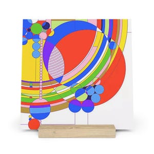 May include: A colorful abstract art print featuring geometric shapes in red, blue, yellow, and green. The artwork is displayed on a white background and supported by a natural wood stand. The design includes circles, arcs, and lines.