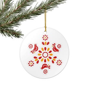 May include: White ceramic Christmas ornament with a folk art design. The ornament features a symmetrical pattern of red birds, flowers, and orange and yellow accents. It hangs from a gold string and is displayed against a green pine branch.