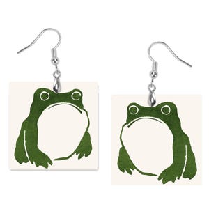 May include: A pair of square earrings featuring a green frog illustration on a cream background. The frog is depicted with a large open mouth and round eyes. The earrings have silver-colored fish hook ear wires.