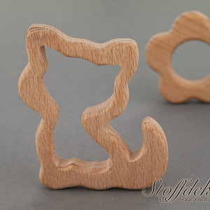 May include: Two wooden teethers for babies. One is shaped like a flower with a hole in the center. The other is shaped like a squiggly line with a hole in the center. Both are made of natural wood.