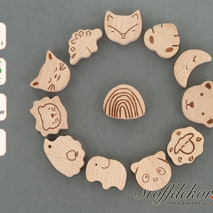 Puede incluir: Un conjunto de 11 mordedores de madera para bebés con varios diseños de animales y formas. Los mordedores están hechos de madera de haya natural y son seguros para los bebés. Los mordedores están dispuestos en círculo sobre un fondo gris. El texto "DIN EN 71-3", "FSC", "Schadstoff Frei" y "Buche" es visible en la imagen.