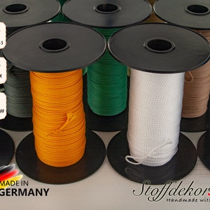 Könnte beinhalten: Spulen mit farbigem Kordel, darunter orange, grün, weiß, beige und blau, auf schwarzen Spulen. Die Spulen sind mit "DIN EN 71-3", "OEKO-TEX 100" und "SCHADSTOFF FREI" gekennzeichnet. Der Text "MADE IN GERMANY" ist ebenfalls sichtbar.