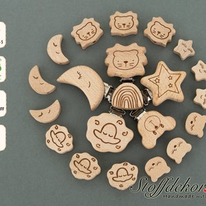 Könnte beinhalten: Ein Set aus Holz-Beißringen für Babys mit verschiedenen Formen und Designs, darunter Monde, Sterne, Wolken und Tiere. Die Beißringe sind aus natürlichem Buchenholz gefertigt und für Babys zum Kauen geeignet. Das Set enthält einen Clip zum Befestigen der Beißringe an einem Kinderwagen oder einer Wickeltasche. Die Beißringe sind handgefertigt und tragen den Text "Stoffdekor.de Handmade with Love".