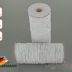 Könnte beinhalten: Zwei Spulen weißen elastischen Kordels, beschriftet mit "DIN EN 71-3", "OEKO-TEX 100" und "SCHADSTOFF FREI". Die Spulen befinden sich auf einem grauen Hintergrund und haben ein Etikett mit der Aufschrift "MADE IN GERMANY" und "Stoffdekor.de Handmade with Love".