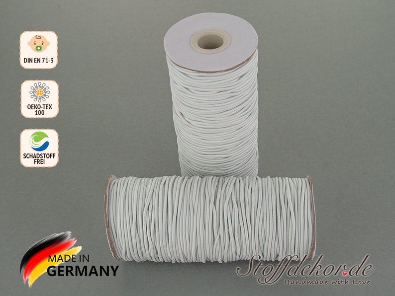 Pu&ograve; includere: Due bobine di cordoncino elastico bianco, etichettate con "DIN EN 71-3", "OEKO-TEX 100" e "SCHADSTOFF FREI". Le bobine sono su uno sfondo grigio e hanno un'etichetta che dice "MADE IN GERMANY" e "Stoffdekor.de Handmade with Love".