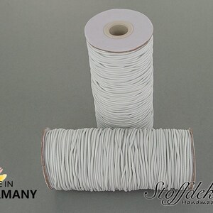 Pu&ograve; includere: Due bobine di cordoncino elastico bianco, ciascuna avvolta attorno a un tubo di cartone. Le bobine sono etichettate con "DIN EN 71-3", "OEKO-TEX 100", "SCHADSTOFF FREI" e "MADE IN GERMANY".