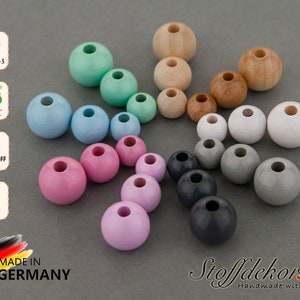 Puede incluir: Una variedad de cuentas de madera en diferentes colores, incluyendo azul claro, rosa, morado, gris y marrón. Las cuentas están dispuestas en un patrón circular sobre una superficie gris. Las cuentas tienen aproximadamente 2,5 cm de diámetro. El texto "DIN EN 71-3", "FSC", "Schadstoff Frei", "Ahorn", "Made in Germany" y "Stoffdekor.de Handmade with Love" es visible en la imagen.