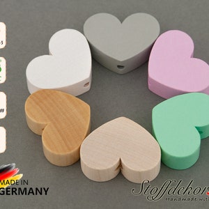 Puede incluir: Seis cuentas de madera en forma de corazón en varios colores, incluyendo blanco, gris, rosa, madera natural, beige y verde. Las cuentas tienen un agujero en el centro para enhebrar. Las cuentas están hechas de madera de arce y son seguras para los niños. DIN EN 71-3, FSC, Schadstoff Frei y Ahorn son visibles en la imagen.