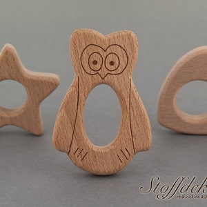 May include: Three wooden teethers in the shapes of a star, an owl, and a heart. The teethers are made of natural beech wood and are safe for babies to chew on. The teethers are labeled with the text "DIN EN 71-3", "FSC", "Schadstoff Frei", and "Buche".