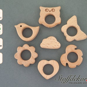 Könnte beinhalten: Satz mit sieben Holz-Beißringen in verschiedenen Formen, darunter ein Eule, ein Vogel, eine Wolke, eine Blume, ein Herz und zwei weitere Formen. Die Beißringe sind aus Naturholz gefertigt und für Babys zum Kauen gedacht.