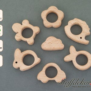 Könnte beinhalten: Satz mit sieben Holz-Beißringen in Form von Blume, Schildkröte, Igel, Wolke, Hase, Fisch und Auto. Die Beißringe sind aus Naturholz gefertigt und für Babys zum Kauen geeignet.