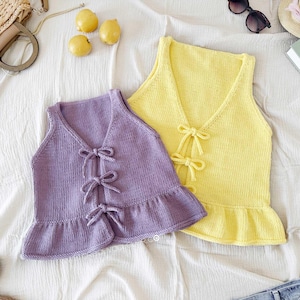 Könnte beinhalten: Zwei gestrickte ärmellose Oberteile, eines in Lavendel, das andere in Gelb. Beide haben einen V-Ausschnitt, Schleifendetails und einen Rüschensaum. Die Oberteile werden auf einer weißen Oberfläche präsentiert. Das Bild enthält auch Zitronen, Sonnenbrillen und einen Strohhut.