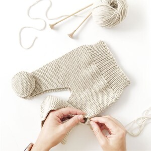 May include: A beige knitted garment with a ribbed waistband and a round pom-pom detail. The item is crafted from light-coloured yarn, accompanied by a ball of matching yarn and two wooden knitting needles positioned above the piece.