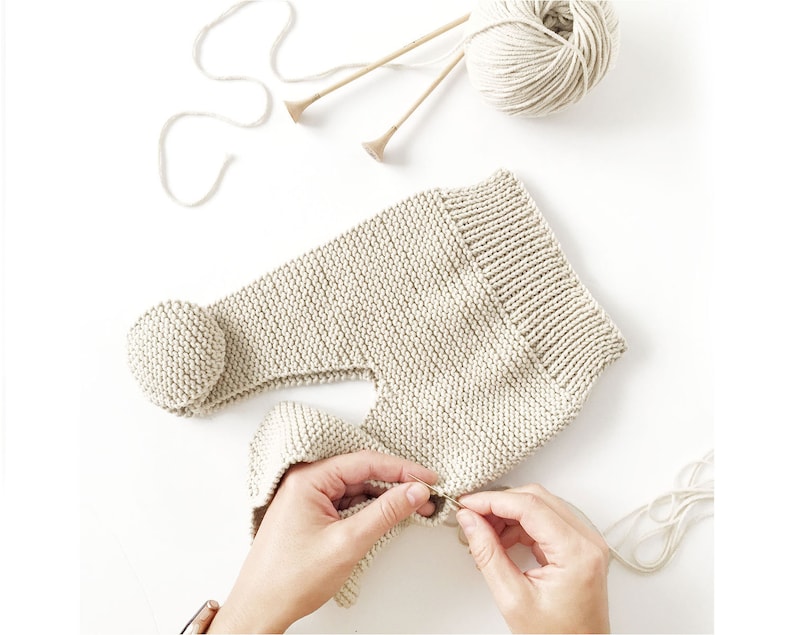 May include: A beige knitted garment with a ribbed waistband and a round pom-pom detail. The item is crafted from light-coloured yarn, accompanied by a ball of matching yarn and two wooden knitting needles positioned above the piece.