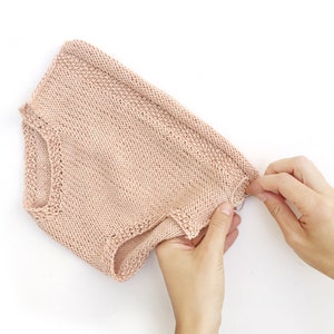 May include: A pale pink knitted baby romper with a delicate lace trim around the leg openings.