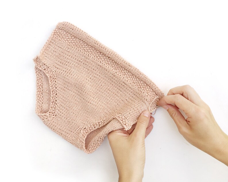 May include: A pale pink knitted baby romper with a delicate lace trim around the leg openings.