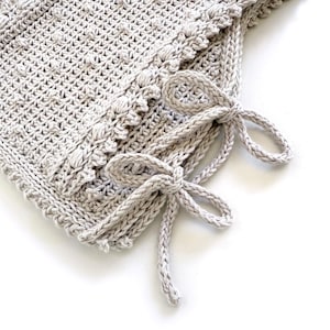 Peut inclure: Gros plan sur une couverture crochet&eacute;e gris clair avec un motif textur&eacute; et une bordure d&eacute;corative. La couverture comporte deux n&oelig;uds crochet&eacute;s nou&eacute;s sur le c&ocirc;t&eacute;. La couverture faite &agrave; la main est parfaite pour un b&eacute;b&eacute; ou comme pi&egrave;ce d&eacute;corative.