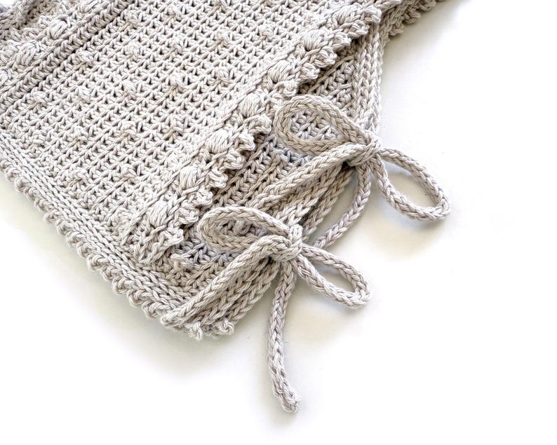 Peut inclure: Gros plan sur une couverture crochet&eacute;e gris clair avec un motif textur&eacute; et une bordure d&eacute;corative. La couverture comporte deux n&oelig;uds crochet&eacute;s nou&eacute;s sur le c&ocirc;t&eacute;. La couverture faite &agrave; la main est parfaite pour un b&eacute;b&eacute; ou comme pi&egrave;ce d&eacute;corative.