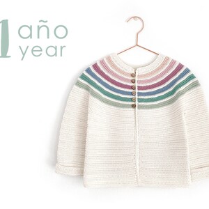 May include: A white crocheted cardigan with a rainbow stripe pattern across the shoulders. The cardigan has a button closure and long sleeves. The text "1 año year" is visible in the background.