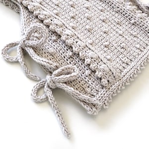 Peut inclure: Un article au crochet fait &agrave; la main, de couleur gris clair, avec un motif textur&eacute; et une bordure d&eacute;corative. L'article comporte deux ensembles de liens, chacun nou&eacute; en n&oelig;ud. Le crochet est d&eacute;taill&eacute; et semble doux et confortable.