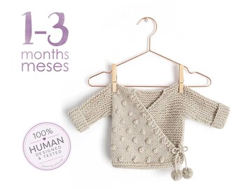 May include: A beige knit wrap sweater for 1-3 months with long sleeves, a textured bobble pattern on the front, and side tie closures with small pom-poms. The garment hangs on a copper-coloured hanger with wooden clips. Text reads 1-3 months meses and 100% human designed & tested.