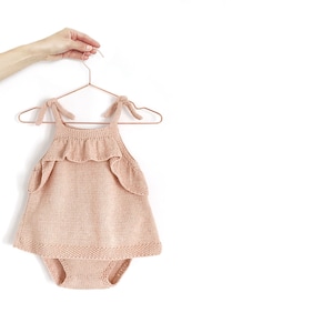 May include: A peach-colored knitted baby onesie with a ruffled neckline and straps. The onesie is hanging on a copper-colored hanger.