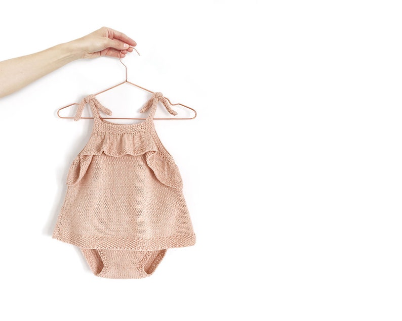 May include: A peach-colored knitted baby onesie with a ruffled neckline and straps. The onesie is hanging on a copper-colored hanger.