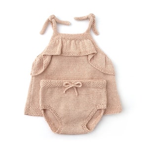 May include: A knitted baby outfit in a light pink color. The outfit includes a sleeveless top with a ruffled neckline and a matching pair of shorts with a bow at the waist.