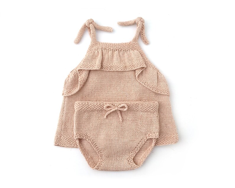 May include: A knitted baby outfit in a light pink color. The outfit includes a sleeveless top with a ruffled neckline and a matching pair of shorts with a bow at the waist.