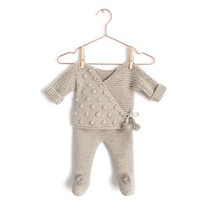 May include: A beige knitted two-piece outfit consisting of a long-sleeved wrap top with a textured bobble pattern and matching footed trousers. The top features a side tie closure with a small pom-pom detail, and the trousers have pom-poms on each foot.