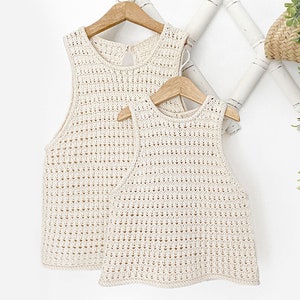 May include: Two crocheted sleeveless tops in a cream color, hanging on wooden hangers against a white background.