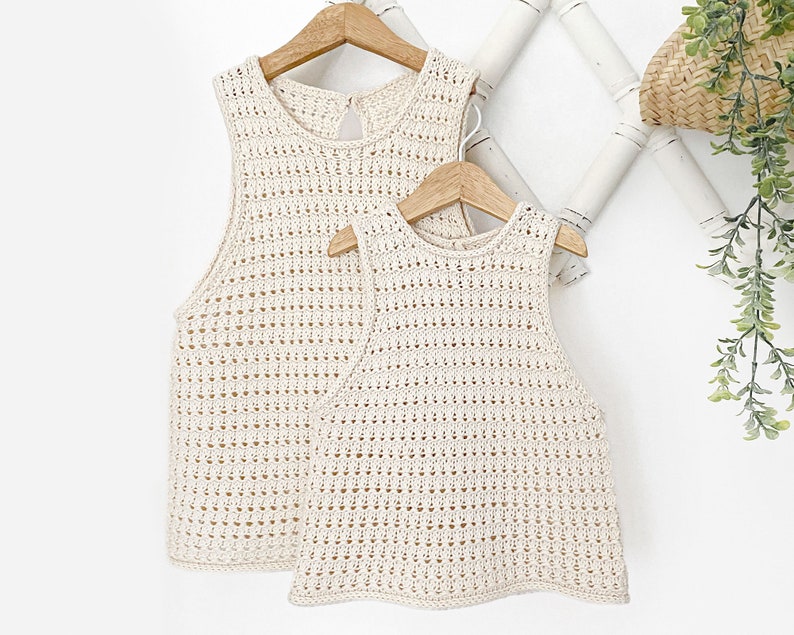 May include: Two crocheted sleeveless tops in a cream color, hanging on wooden hangers against a white background.