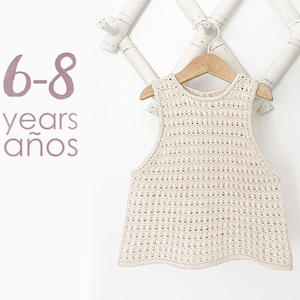 May include: A white crocheted sleeveless top for children, hanging on a wooden hanger. The top has a delicate openwork pattern. The text "6-8 years a&ntilde;os" is visible in the image.
