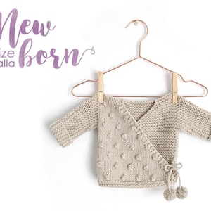 May include: A beige knitted baby wrap with a tie closure and pom-poms. The wrap has a textured pattern with small raised dots. The text "New born" is written in purple above the wrap.