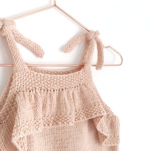 May include: A pale pink knitted tank top with ruffled trim and knotted straps. The top is hanging on a copper-colored hanger.