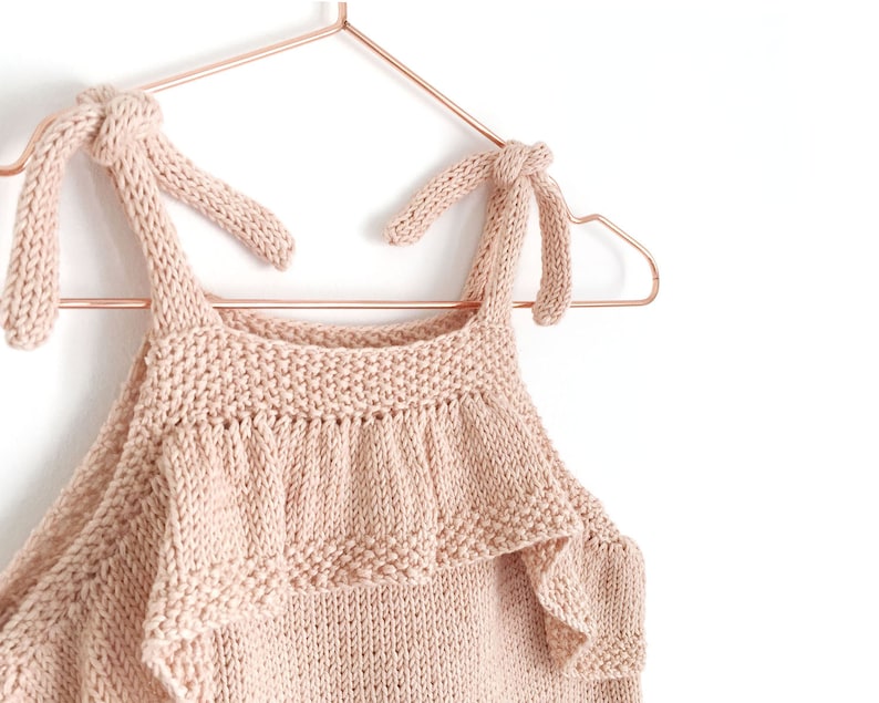 May include: A pale pink knitted tank top with ruffled trim and knotted straps. The top is hanging on a copper-colored hanger.