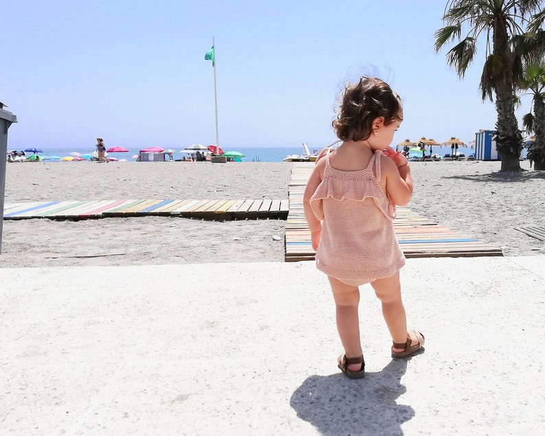 Op de afbeelding: Een jong kind in een roze gehaakt jurkje en sandalen loopt op een strandpromenade. Het kind is met de rug naar de camera gekeerd en kijkt naar rechts. Het strand is op de achtergrond.