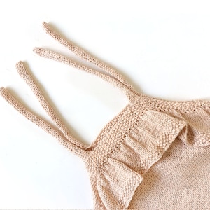 May include: A light pink knitted baby romper with ruffled straps. The romper is made of a soft, lightweight material and has a relaxed fit.