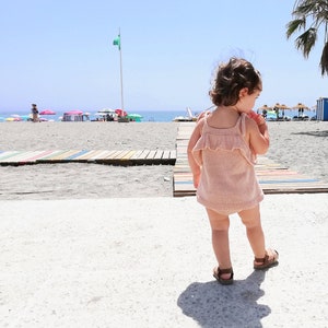 May include: A young child wearing a pink crocheted dress and sandals walks on a sandy beach with a colorful wooden walkway in the background.