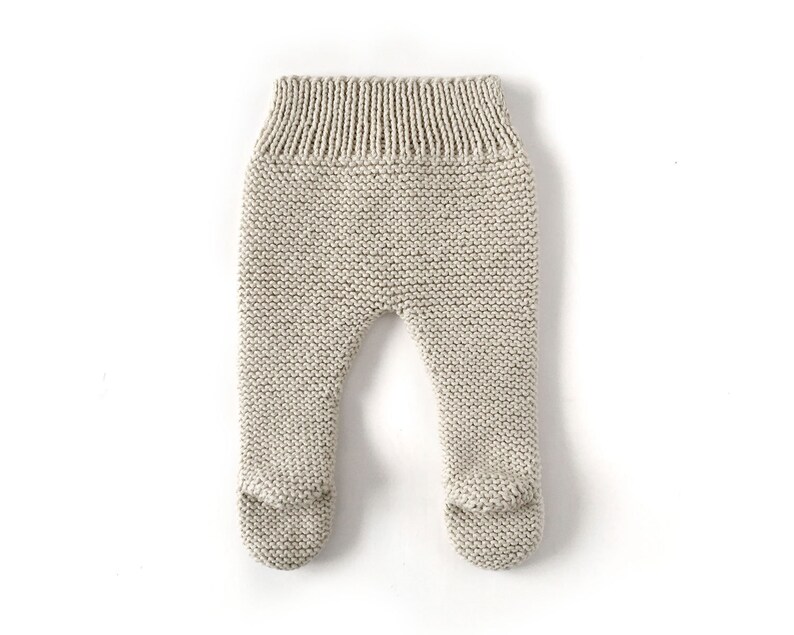 May include: Beige knit footed trousers featuring a garter stitch texture throughout the legs and a ribbed elastic waistband. The garment is constructed with integrated foot coverings at the base of each leg, providing a seamless design.