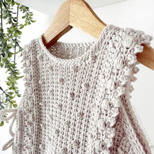 Peut inclure: Un haut sans manches en crochet beige clair avec un motif textur&eacute; et des bordures d&eacute;coratives, suspendu &agrave; un cintre en bois. Le haut pr&eacute;sente une encolure ronde et des d&eacute;tails de liens sur les c&ocirc;t&eacute;s. Le crochet a un motif de point de bouff&eacute;e.