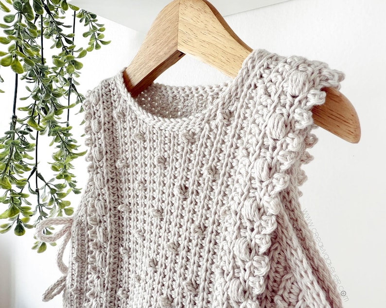 Peut inclure: Un haut sans manches en crochet beige clair avec un motif textur&eacute; et des bordures d&eacute;coratives, suspendu &agrave; un cintre en bois. Le haut pr&eacute;sente une encolure ronde et des d&eacute;tails de liens sur les c&ocirc;t&eacute;s. Le crochet a un motif de point de bouff&eacute;e.
