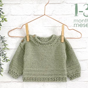 Op de afbeelding: Een saliegroene gebreide babytrui met houten knopen die aan een koperen hanger hangt. De trui is voor baby's van 1-3 maanden oud.