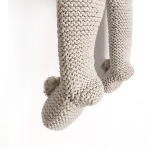 May include: A pair of light beige knitted leggings featuring a textured garter stitch pattern. Each leg ends in a footed design with a small, matching beige pom-pom attached to the heel area of the foot, creating a soft and uniform appearance.