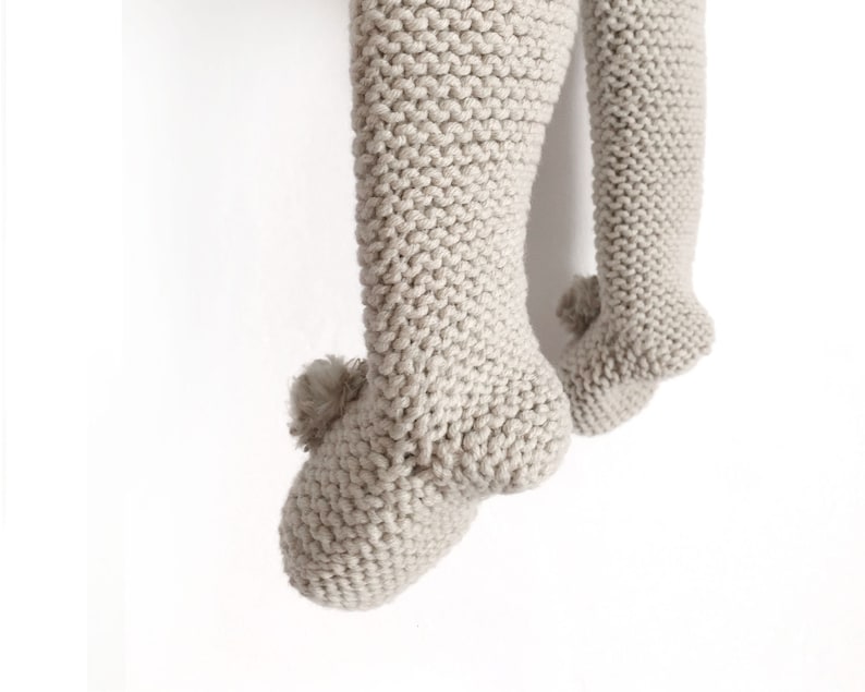 May include: A pair of light beige knitted leggings featuring a textured garter stitch pattern. Each leg ends in a footed design with a small, matching beige pom-pom attached to the heel area of the foot, creating a soft and uniform appearance.