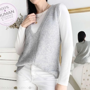 May include: A light gray sleeveless knit vest with a deep V-neckline, worn over a long-sleeved white shirt and white pants. A circular badge in the upper left corner displays the text 100% HUMAN DESIGNED & TESTED CREATIVE ATELIER.