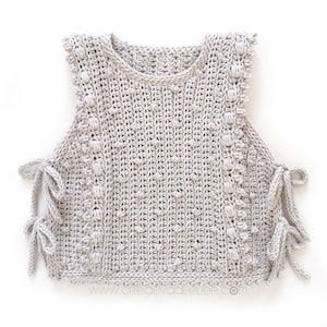 Peut inclure: Un haut sans manches en crochet gris clair, avec un col rond et des liens lat&eacute;raux. Le haut pr&eacute;sente un motif de point textur&eacute; avec des d&eacute;tails en forme de boules sur les bords. L'ourlet du bas est &eacute;galement crochet&eacute;. Le site web www.creativatelier.com est visible.