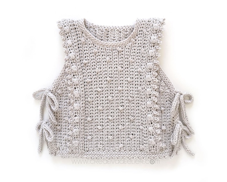 Peut inclure: Un haut sans manches en crochet gris clair, avec un col rond et des liens lat&eacute;raux. Le haut pr&eacute;sente un motif de point textur&eacute; avec des d&eacute;tails en forme de boules sur les bords. L'ourlet du bas est &eacute;galement crochet&eacute;. Le site web www.creativatelier.com est visible.
