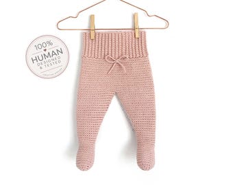 Puede incluir: Pantalones con pies de ganchillo en color rosa empolvado, con cintura alta acanalada y un lazo decorativo en el centro. La prenda cuelga de una percha de alambre color cobre. Una insignia circular a la izquierda muestra el texto 100% HUMAN DESIGNED & TESTED.