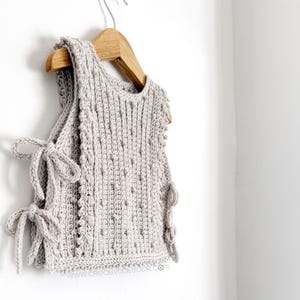 Peut inclure: Un haut sans manches en crochet gris clair avec un motif textur&eacute; et des liens lat&eacute;raux. Le haut est suspendu &agrave; un cintre en bois contre un mur blanc. Les liens lat&eacute;raux sont nou&eacute;s en n&oelig;uds.
